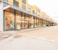 Multi-story commercial building with a large retail store called Fust occupying the ground floor. The building has a modern, glass-fronted design with balconies on the upper floors. There is a paved area in front of the building that appears to be used fo