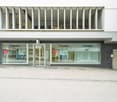 The Audika building is a white, modern structure with large glass windows, a brick wall, and white shutters.