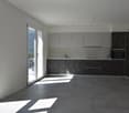 Spacious open-plan kitchen with modern, minimalist design. Gray cabinets, black marble countertops, and large windows providing natural light. Sliding glass door leading to a balcony or terrace.