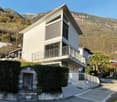 Modern 2-story house, white facade, balcony with metal railing, driveway with concrete curb, lush greenery, hilly background, clear sky