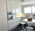 Modern, minimalist kitchen with white cabinets, stainless steel appliances including a dishwasher, white countertops, and dark tile flooring. The kitchen has a breakfast bar with white stools and a window providing natural light.