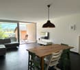 Spacious open-plan living area with a large sofa, wooden dining table and chairs, a TV stand, and a sliding glass door leading to a balcony or terrace with a scenic view of the surrounding landscape.