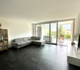 Spacious living room with large windows, grey sectional sofa, bookshelf, and sliding glass doors leading to a balcony or terrace.