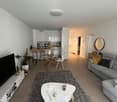 white and beige living room with large gray rug, round table, two sofas, TV on white shelf, open kitchen with white counters