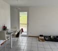 Bright and spacious living room with a dining area, tiled flooring, and a balcony or terrace visible through the window. The room contains a dining table with chairs, a small desk, and some storage boxes and items.