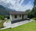 Single story house, white exterior, gray roof, surrounded by lush greenery, stone patio, garden table, distant house and mountain landscape