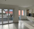 Spacious open-plan living area with large windows and sliding glass doors leading to a balcony or terrace, modern kitchen with white cabinets and appliances, tiled flooring throughout