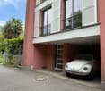 2-story residential building with a red exterior, white windows, and shutters. A classic Volkswagen Beetle is parked in the garage underneath the building.