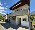 2-story house with wooden roof, stucco exterior, balcony, and paved parking area. The house is surrounded by greenery and mountains in the background.