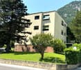 Building, multi-story, beige walls, windows with green shutters, surrounded by green lawn, trees, bushes, mountain in background, cars parked nearby