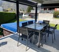 Outdoor dining table with chairs, covered by a retractable roof, surrounded by a garden with a hedge and a grassy lawn, with a view of the mountains