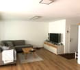 L-shaped grey sofa, wooden coffee table, white TV unit, flat screen TV, floor area rug, brown rug by the door, ceiling mounted lighting