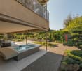 Balcony with a jacuzzi, chairs, and a cozy seating area. The outdoor space includes lush greenery, shrubs, and trees. The background shows a building with a gray exterior.