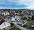 Aerial view of a town with residential buildings, some under construction, a parking lot, and various trees and plants. The houses vary in size and architecture.