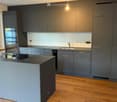 modern kitchen, dark grey cabinets, wooden floor, island with sink, electric kettle, stainless steel fridge, dishwasher, oven