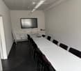 Empty room with white walls, black floor, long white tables, black chairs, mounted TV, whiteboard, radiator