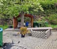 A garden with a wooden gazebo, a table and chairs, a potted plant, a blue barrel, a green tank, a yellow hose, and a stone wall.