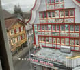 Aerial view of a building with multiple floors, red trim, and many windows. The building has a name board that reads 'BAZAR HERSCHE'. The building has a small garden with a few potted plants. The building is surrounded by other buildings and a street.