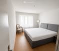 Minimalist bedroom with a single bed, a wooden chair, and a lamp near the window. The room features light wooden flooring and white walls.