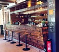 Wooden bar counter with hanging wine glasses, bottles, and stools, illuminated by hanging lamps.