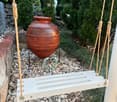 Garden / Outdoor Area. Garden area with a large terracotta vase on a stone base, surrounded by rocks and greenery, with a hanging white bench or swing in the foreground.