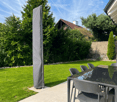 Garden / Outdoor Area. Well-maintained garden with a neatly trimmed lawn, tall trees, and a patio area featuring a glass-top dining table and chairs. A tall, grey outdoor umbrella is positioned near the lawn.