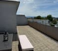 Roof terrace with brick tiles, a small table, two chairs, a bench, and a lantern, overlooking a cityscape with mountains in the background.