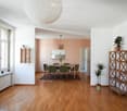 Bright, open-plan living and dining area with wooden flooring, large windows, a round pendant light, a dining table with six chairs, shelves with plants, and a small kitchen area with cabinets and a mirror.