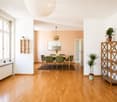 Bright, open-plan living and dining area with wooden flooring, large windows, and modern furniture including a dining table, chairs, shelves, and potted plants.