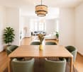 Bright, open-plan living and dining area with wooden flooring, large windows, a dining table with six chairs, a sofa, an ottoman, a floor lamp, and decorative plants.