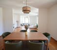 Bright, open living and dining area with wooden flooring, large windows, a dining table with six chairs, a sofa, an ottoman, a floor lamp, and decorative vases.