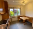 Compact kitchen with wooden cabinets, an induction stove, a small dining table with bench seating, and large windows overlooking a garden.