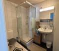 Modern bathroom with a glass-enclosed shower, toilet, sink, and mirror. Includes a small wooden cabinet, towels, and various toiletries.
