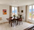 Bright dining area with a wooden table, chairs, vases with flowers, artwork, and a view of mountains through large windows.