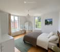 Bright bedroom with wooden flooring, large windows, bed with pillows and blanket, small stool, mirror, painting, and a small table with chair.