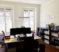 Office space with a large desk, dual monitors, chairs, shelves filled with books and files, and a fan. Natural light from large windows.