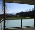 Balcony with glass railing, overlooking a green field, trees, and distant mountains.