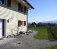 Front garden area with a paved pathway leading to the house entrance, small plants, a wooden fence, and a scenic mountain view in the background.