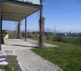 A paved pathway leads to a covered balcony area with a view of a grassy lawn, distant fields, and mountains. Small purple flowers are planted along the pathway.