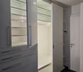 Modern, empty walk-in closet with built-in storage units, glass-fronted shelves, and drawers. The space features sleek, grey cabinetry with metallic handles and a dark tiled floor.