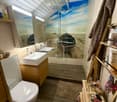 Modern bathroom with a large shower area featuring a beach-themed mural, a white sink, a toilet, and a wooden shelf holding towels and toiletries.
