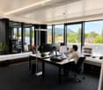 Modern office space with large windows, a desk with multiple monitors, a laptop, and a comfortable chair. The room features a sleek design with a black carpet, white walls, and a view of greenery outside.