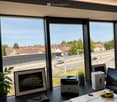 Modern office space with large windows overlooking a cityscape, featuring a desk with a printer, screen, laptop, cellphone, picture frame, plant, and a small toy car.