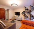 Modern living room with a grey sofa, ottoman, coffee table, TV stand, and wall-mounted shelves. The room features wooden flooring and a large round ceiling light.