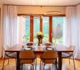 Bright dining room with large windows offering a scenic view, a wooden dining table set with plates, glasses, and a vase of flowers, surrounded by modern chairs.