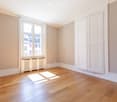 Empty room with wooden flooring, large window, radiator, and white walls.