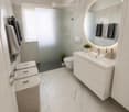Modern bathroom with marble tiles, white sink, large round mirror, toilet, and wicker baskets.