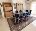 Dining room with a glass table, six blue chairs, a large patterned rug, and wooden cabinets with glass doors.
