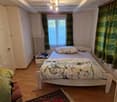 A small bedroom with a single bed, a nightstand, a chair, and a radiator. The room features wooden flooring and a window with green curtains.