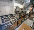 Commercial kitchen with stainless steel appliances including a gas stove, griddle, and oven. Shelves hold plates and utensils, with a cardboard box on the floor.